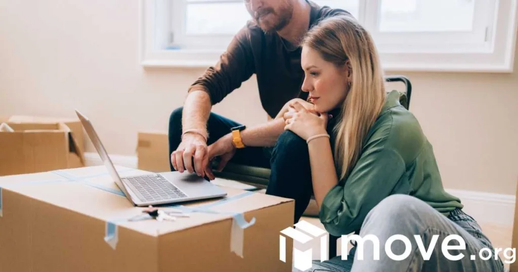 A couple looks at online moving quotes on a laptop, surrounded by boxes.
