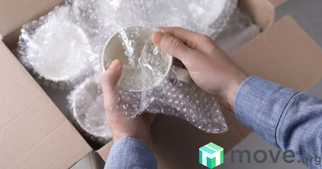 Hands wrapping a mug with bubble wrap above a box filled with wrapped dishes