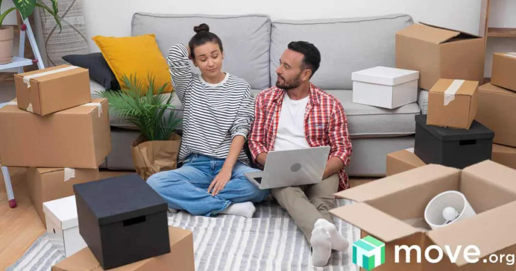 Couple of young people moving into new modern apartment setting up new internet while sitting on floor near sofa among cardboard boxes.